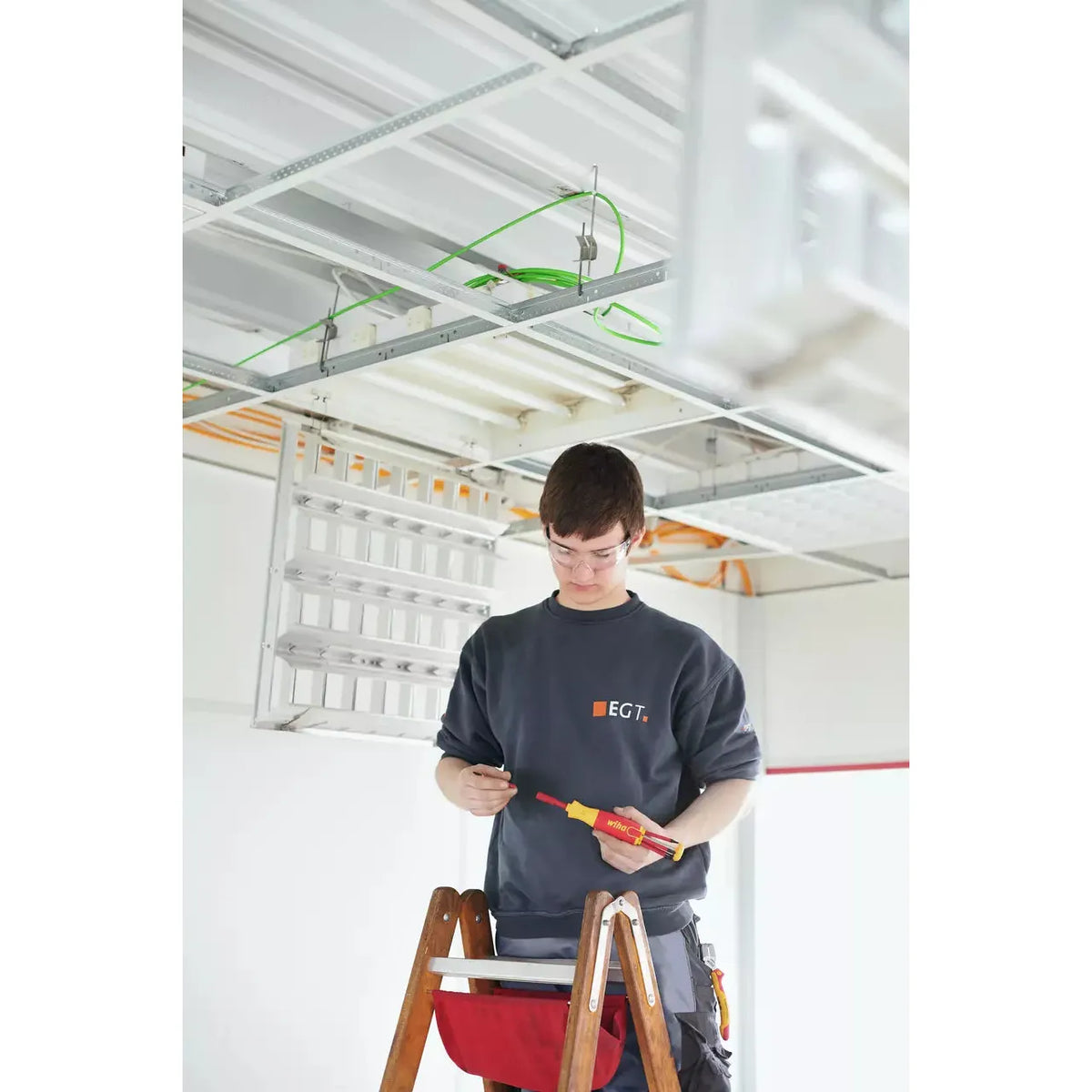 A person on a ladder working with an electric screwdriver near a ceiling with exposed wiring.
