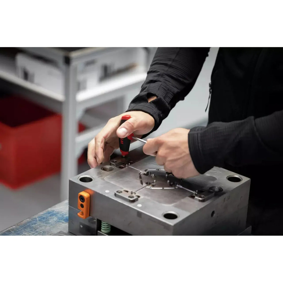 A person using a red-handled screwdriver on a metal mechanical part.