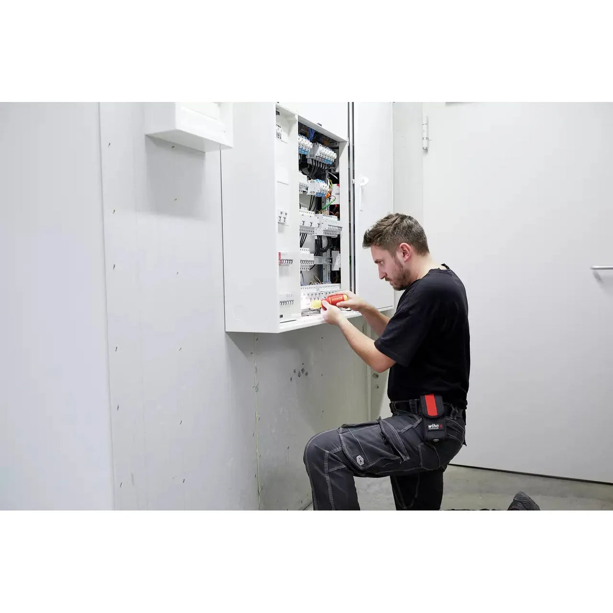 A person kneels while using an electric screwdriver on an open electrical panel