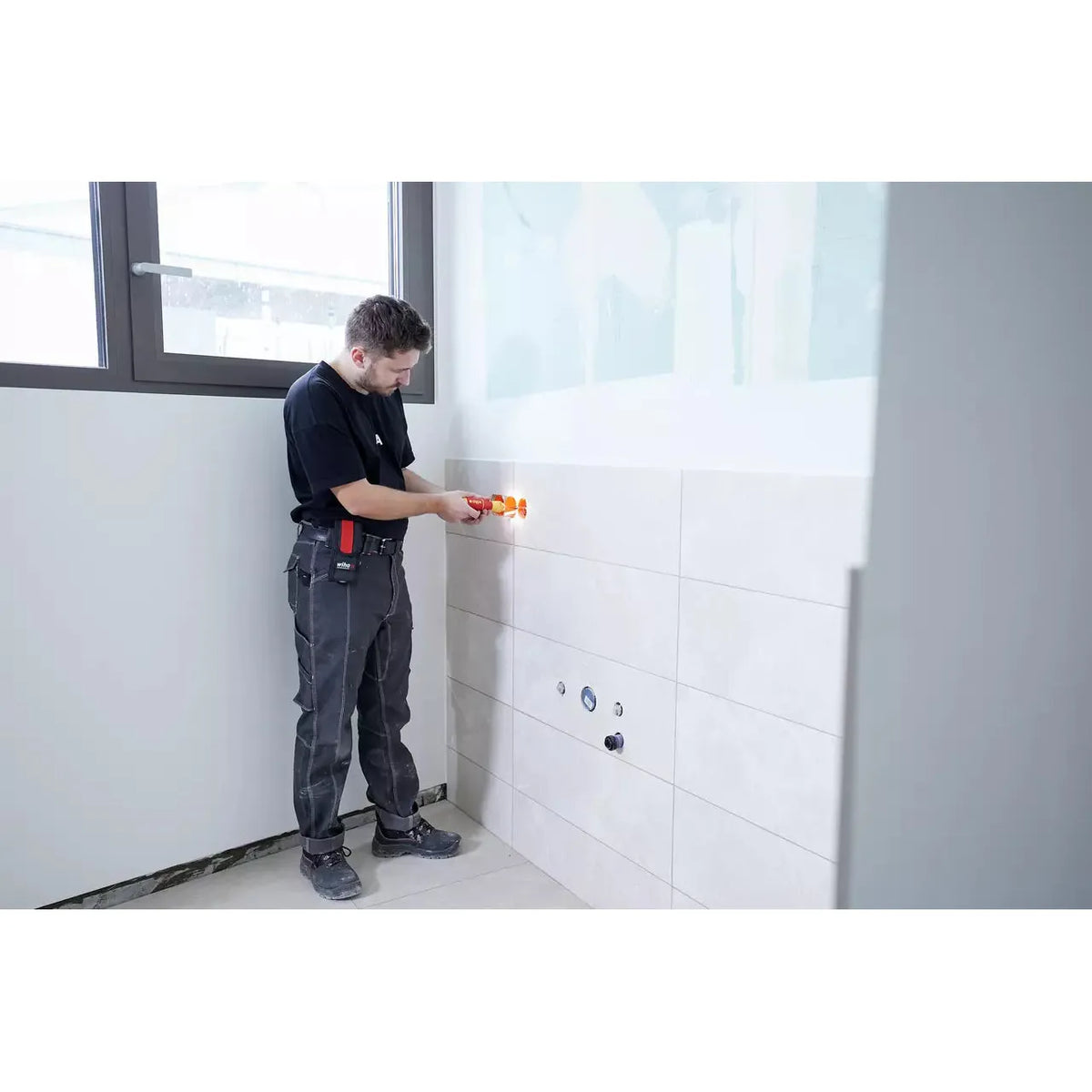 A person using an electric screwdriver on a tiled wall in a bright room.