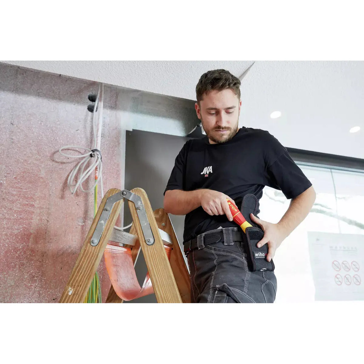 A person on a ladder holds a red and yellow electrical tool in a holster. Wires are visible in the background.