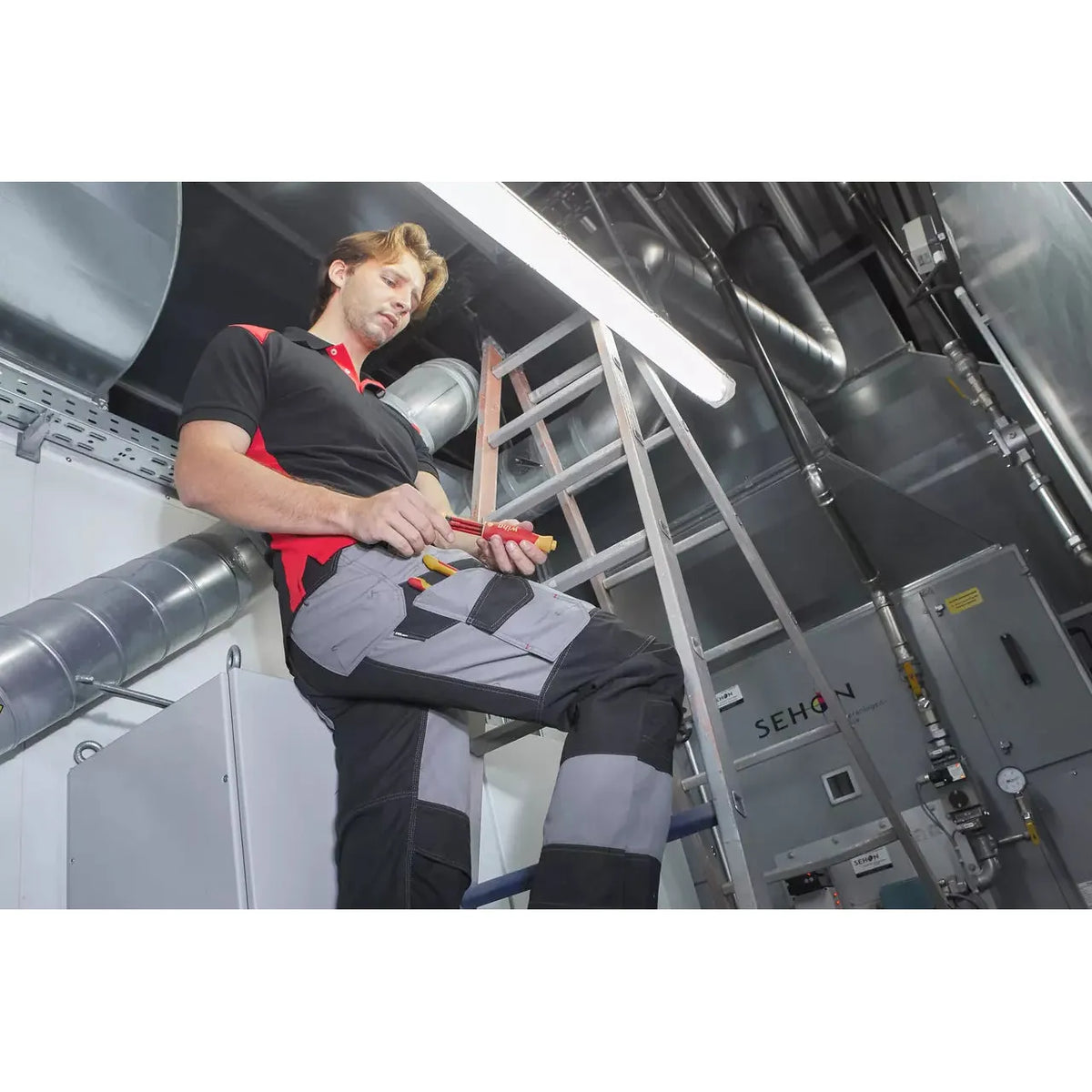 A person working in an industrial setting, holding a tool, with ducts and a ladder in the background.