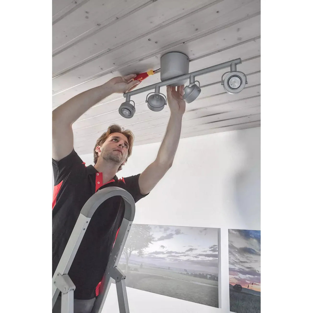 A person on a ladder installs a ceiling light using a screwdriver.