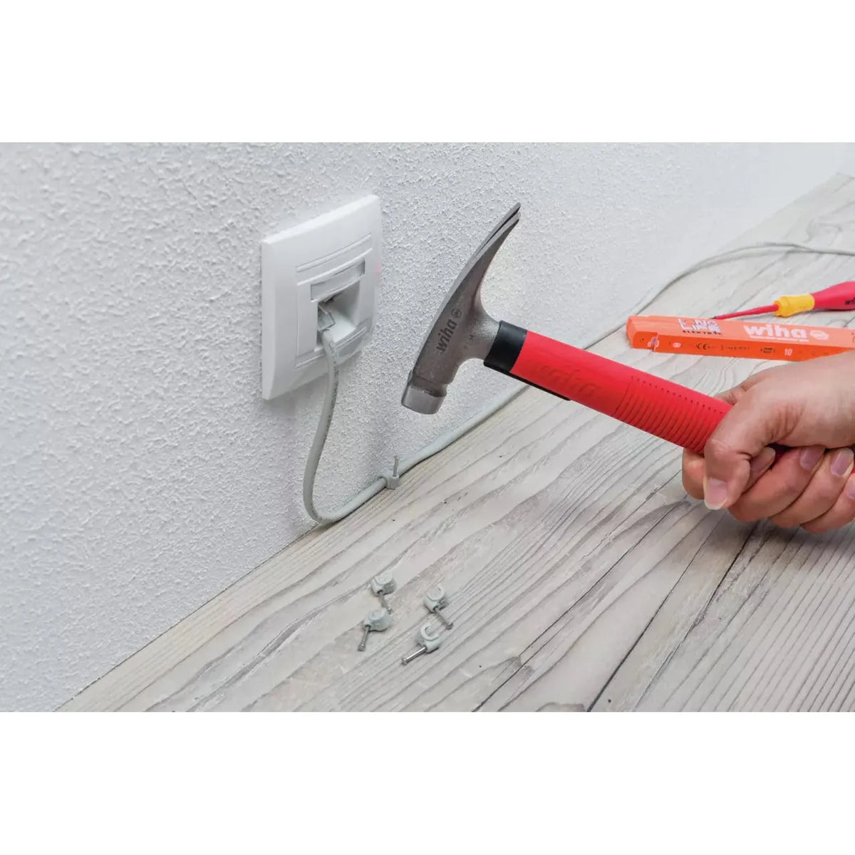 Electrician's hammer with red handle used to secure cable clips on a wooden surface near an outlet.