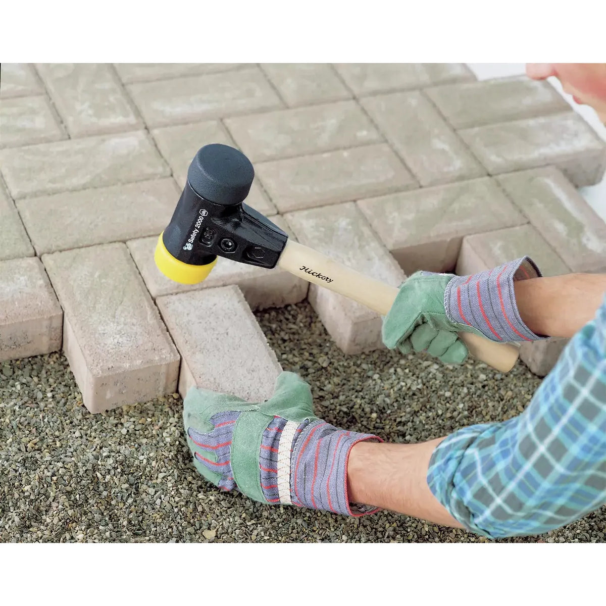 A person using a rubber mallet to adjust paving stones on gravel. The mallet has a wooden handle and black head with a yellow tip.