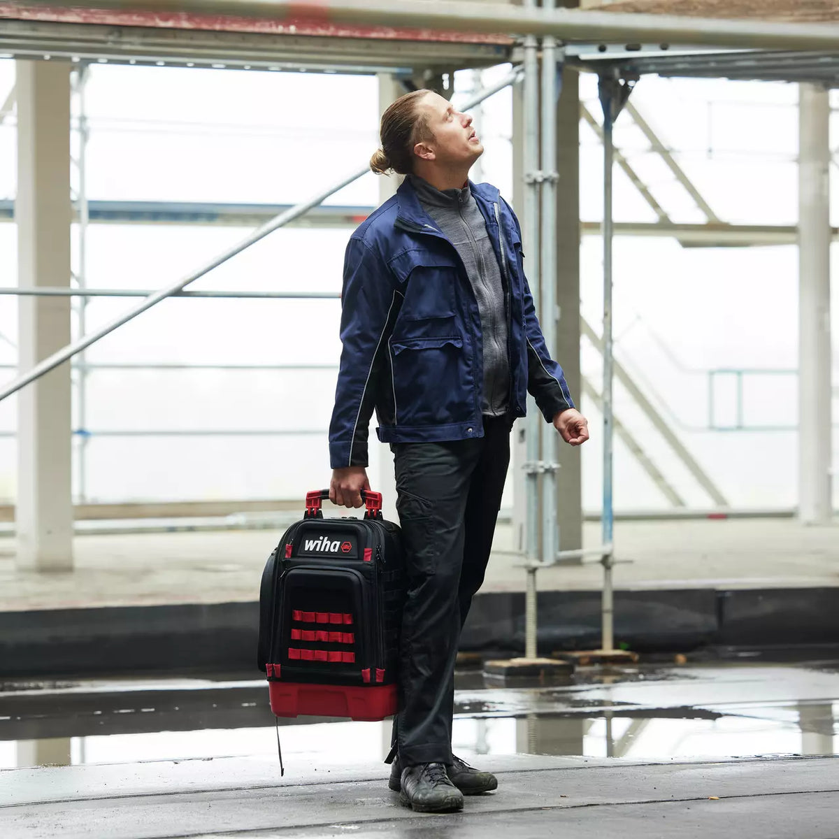 Person in workwear carrying a tool case and looking upwards, construction site environment.