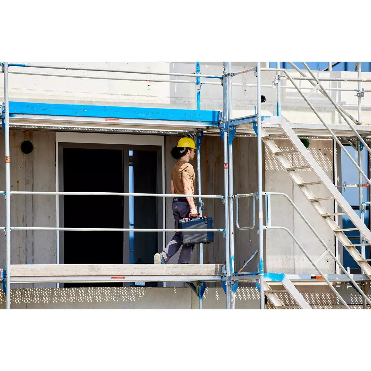 A person in a yellow hard hat carrying a toolbox on a construction site scaffold.
