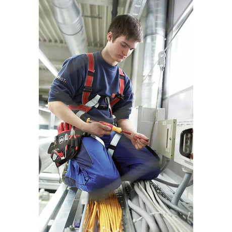 A person is using an electric screwdriver while kneeling on a metal structure with cables. They are wearing safety gear.