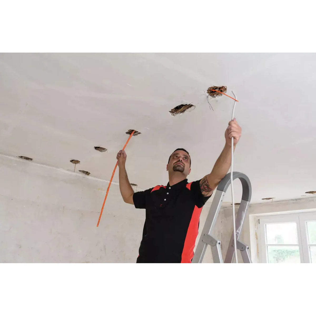A person works on a ceiling, holding a measuring tape and a cable