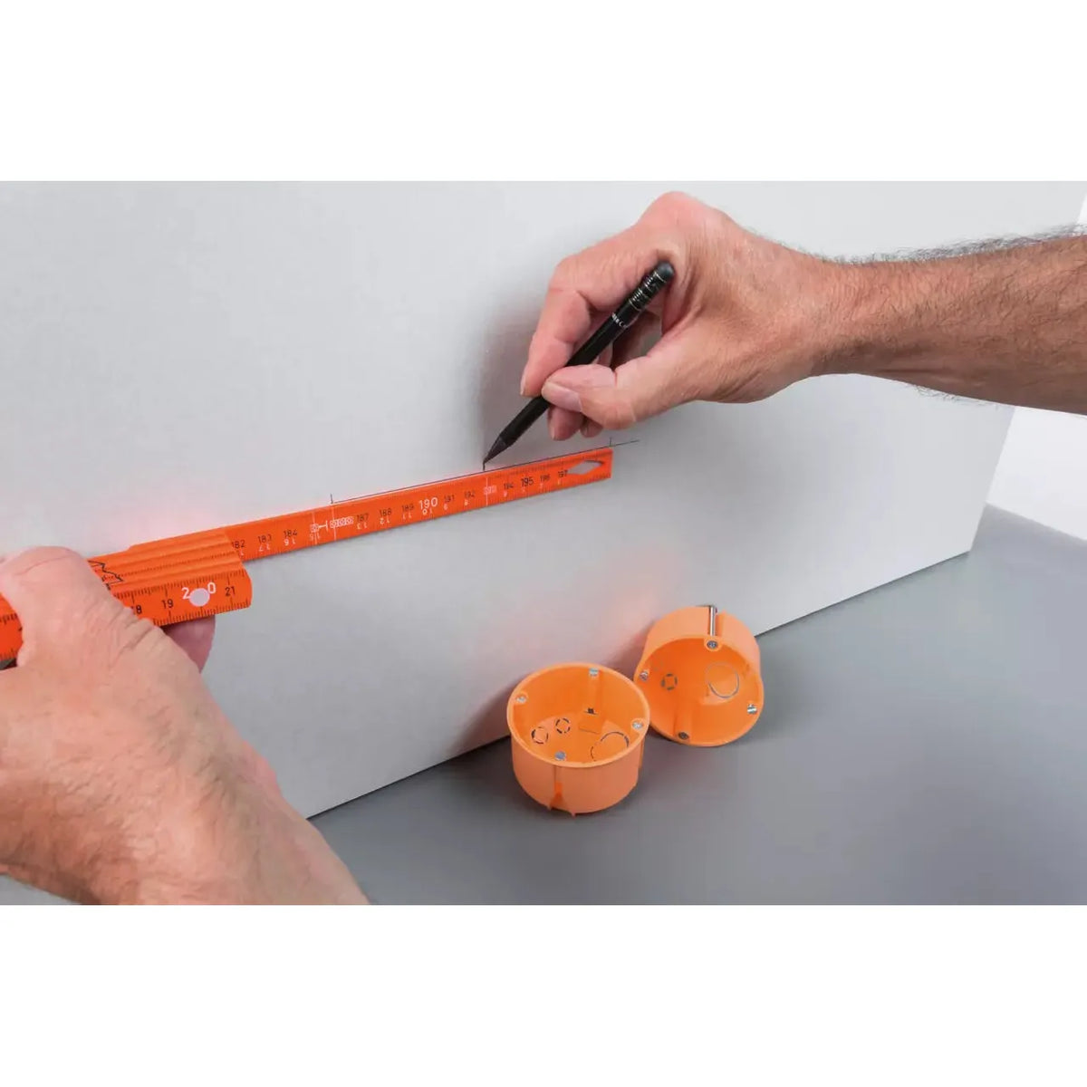 Person using an orange folding ruler to measure and mark on a white surface, with orange electrical boxes nearby.