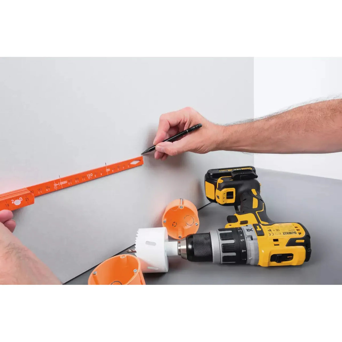 Person using a folding ruler and pencil on a wall, near a drill and orange boxes.
