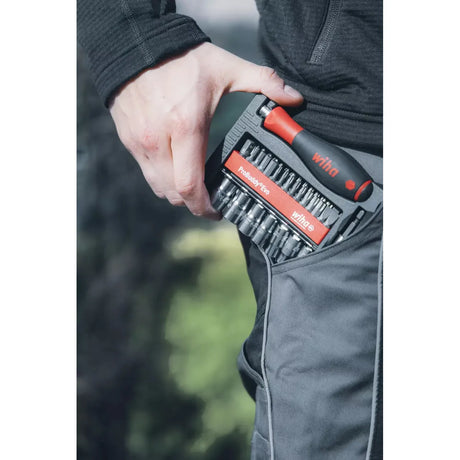 A person holds a tool set with a red and black handle and multiple bits in a trouser pocket.