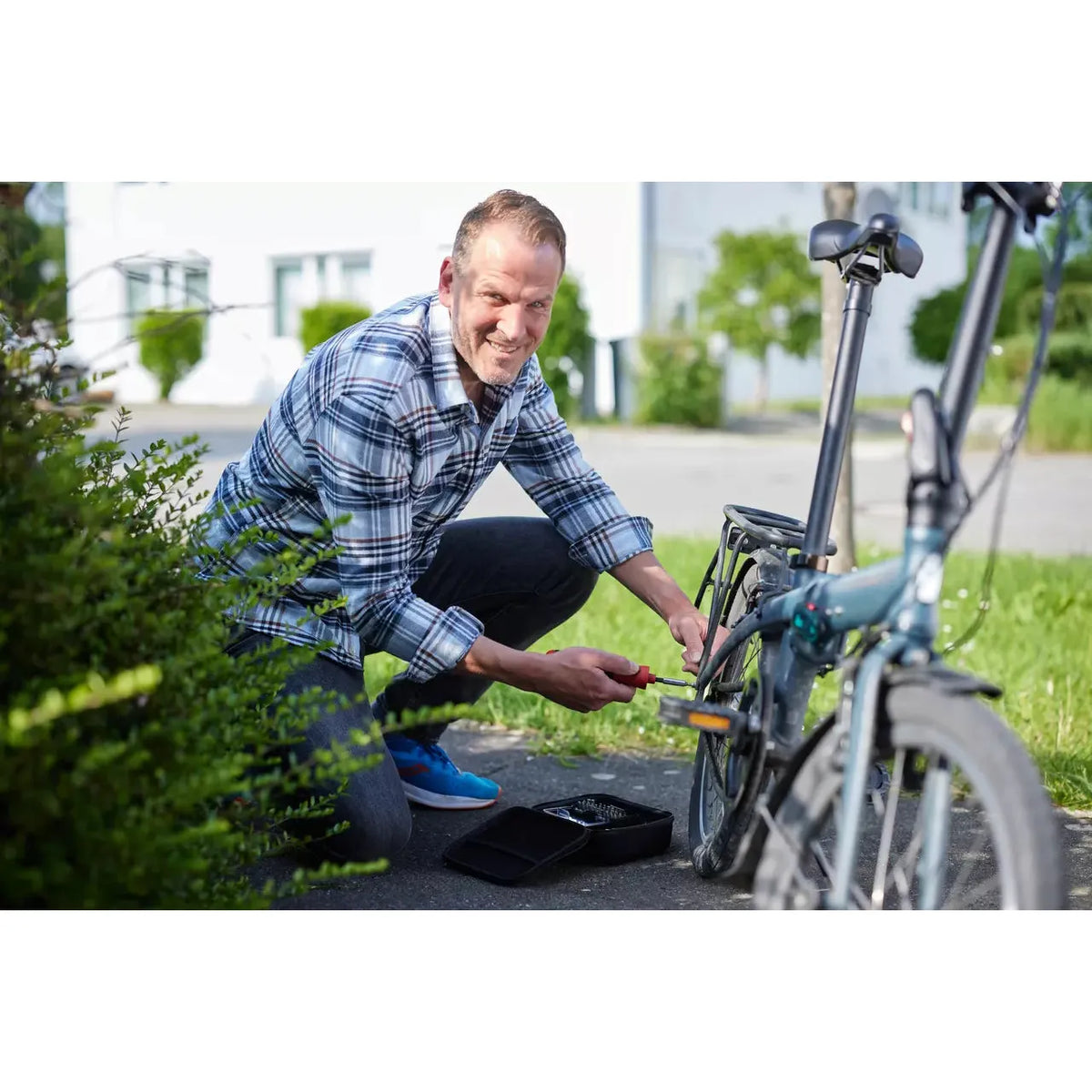 A person kneels outside, using a screwdriver to repair a bicycle. Tool kit is open on the ground.