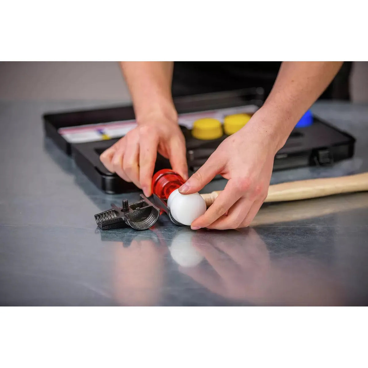 A person assembling a red and white mallet head on a wooden handle next to a toolbox on a metal surface.