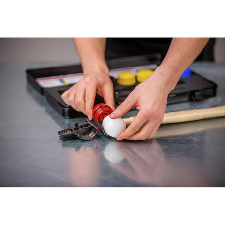 A person assembling a red and white mallet head on a wooden handle next to a toolbox on a metal surface.