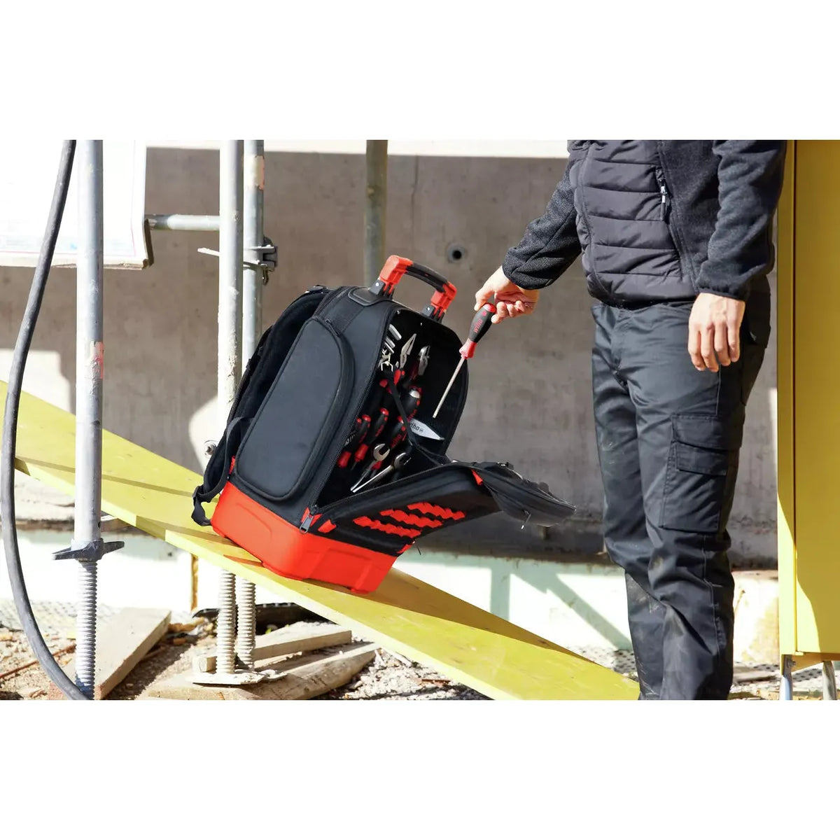 Person taking a screwdriver from a black and red tool backpack on a construction site.