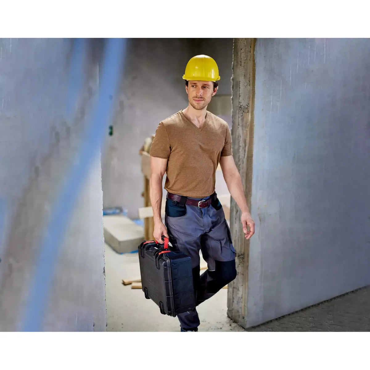 A person in a yellow hard hat carrying a black toolbox in a construction site.