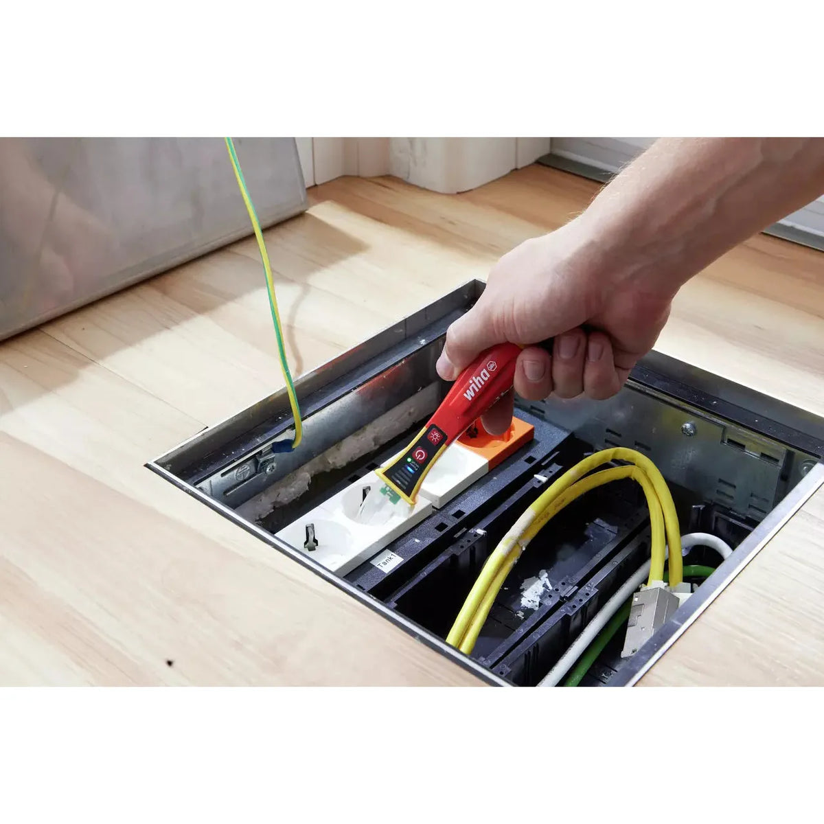 12–1,000V AC – A person using a red non-contact voltage detector near a power socket in a floor box.