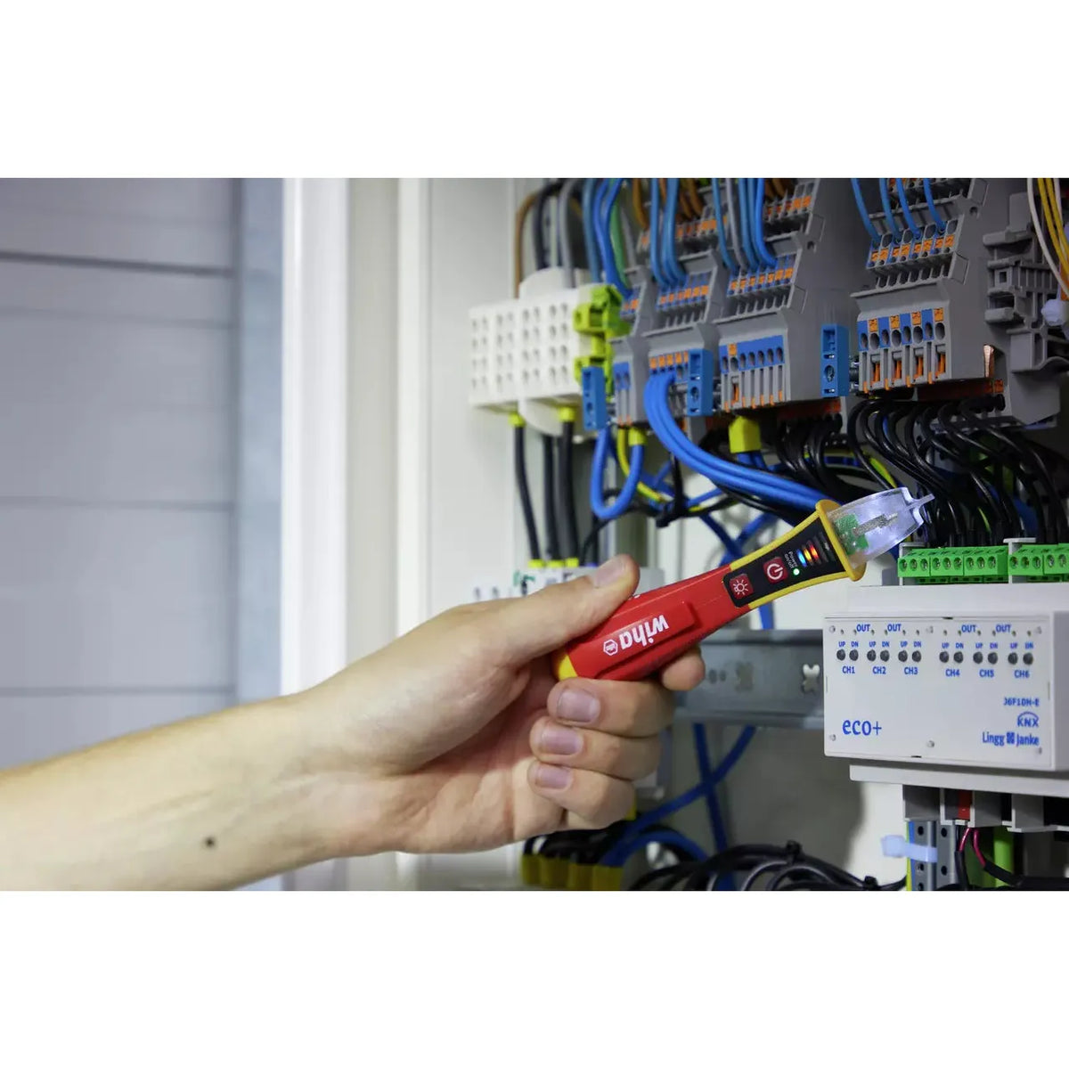 90–1,000V AC – Hand holding a red non-contact voltage detector near wires in an electrical cabinet.