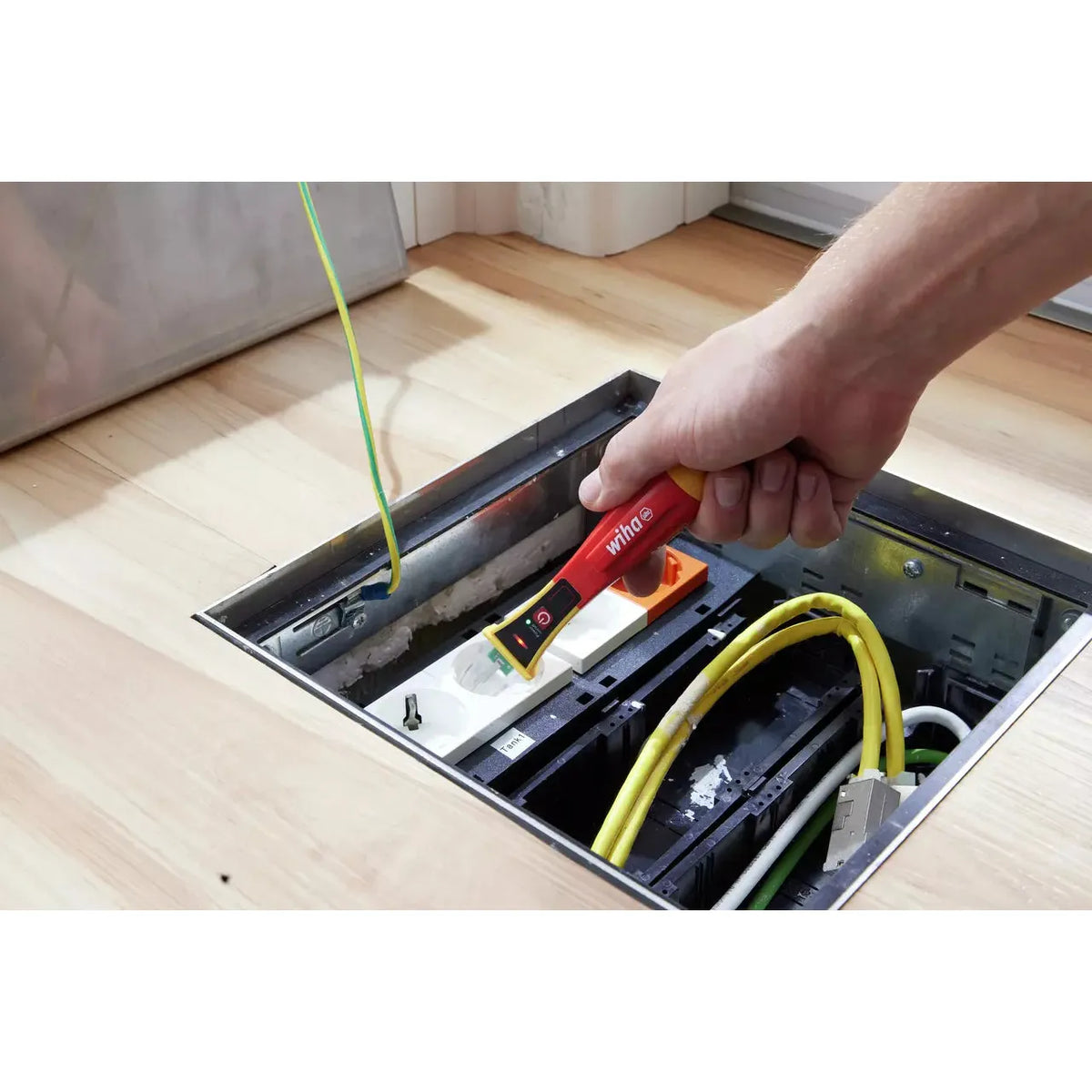 90–1,000V AC – A person using a red non-contact voltage detector on a floor socket with cables