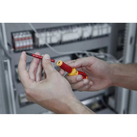 Person holding a red and yellow insulated screwdriver handle and blade in front of electrical panel.