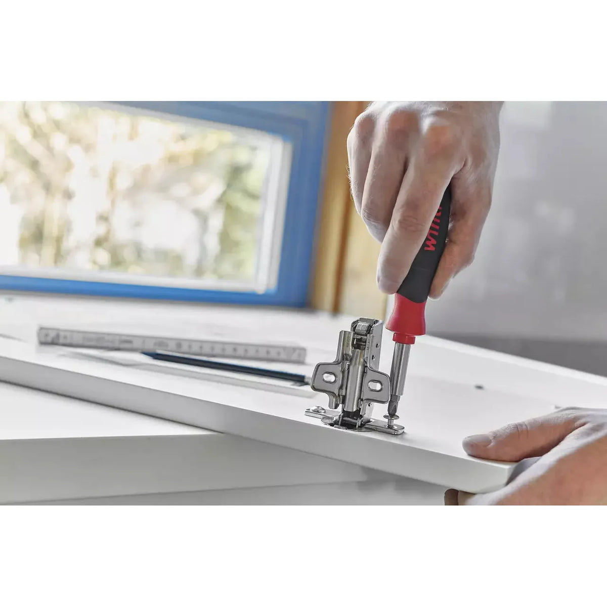 Person using a screwdriver to attach a hinge to a white cabinet door in a kitchen.