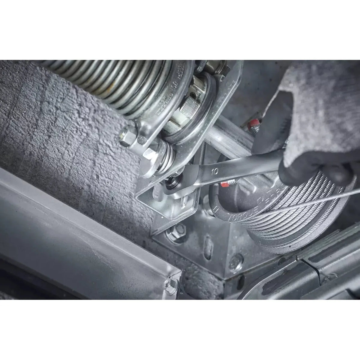 A gloved hand uses a spanner to tighten a nut on a metal mechanism with cables and springs.