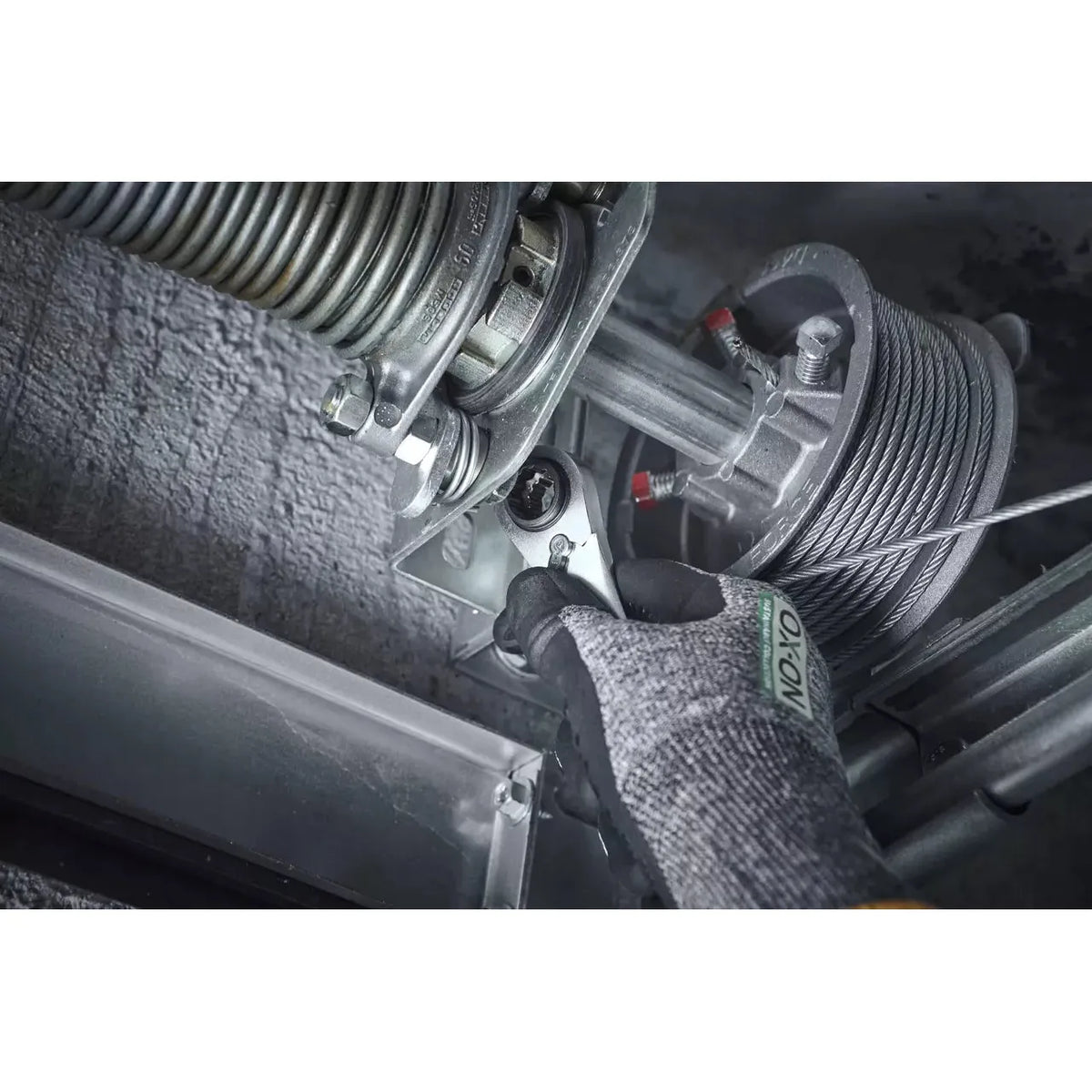 A person uses a ratchet wrench on a metal mechanism with a coiled spring and cable. They are wearing gloves.