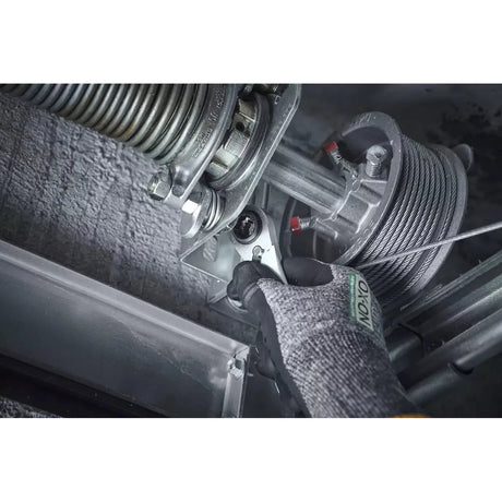 A person uses a ratchet wrench on a metal mechanism with a coiled spring and cable. They are wearing gloves.