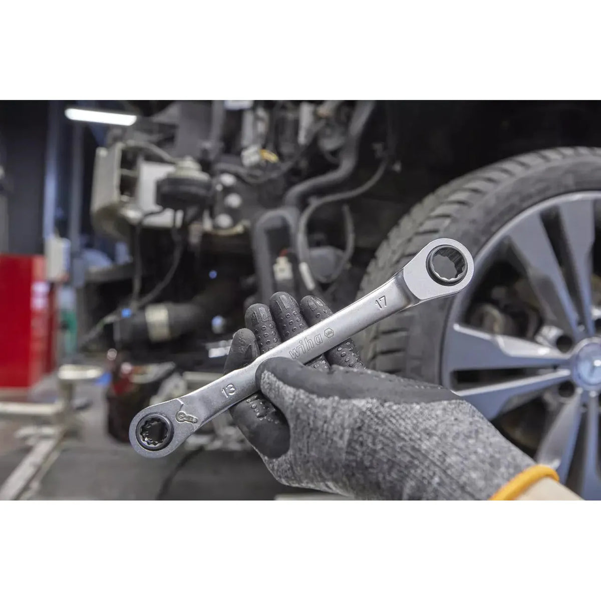 A gloved hand holding a double-ended ratchet wrench in front of a car wheel.