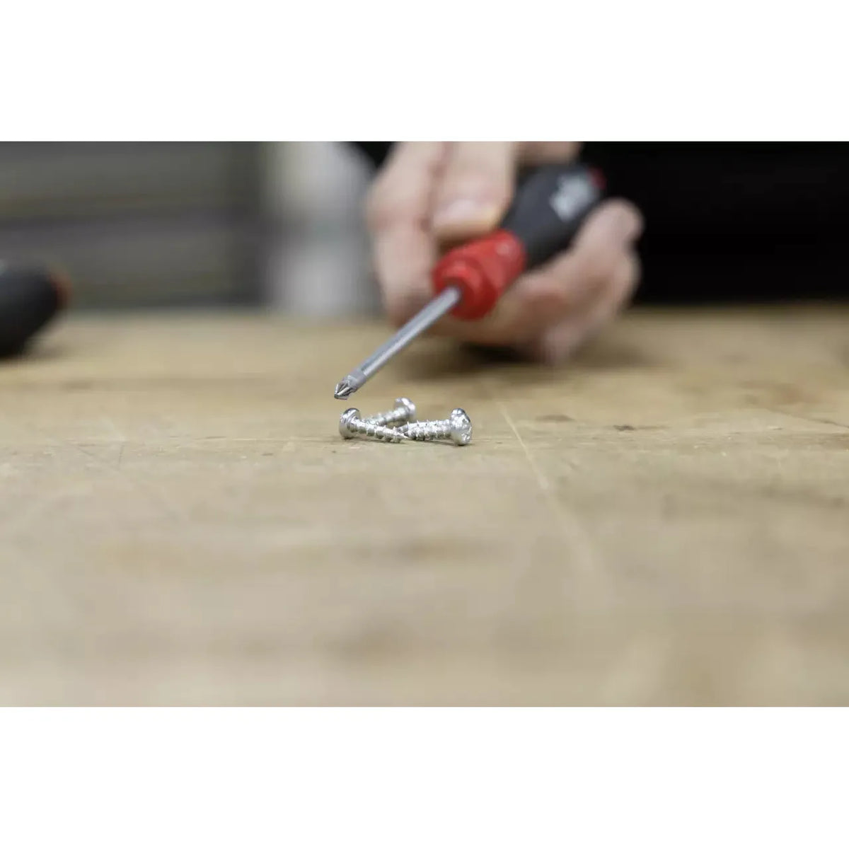 A person holding a red-handled screwdriver near three screws on a wooden surface.