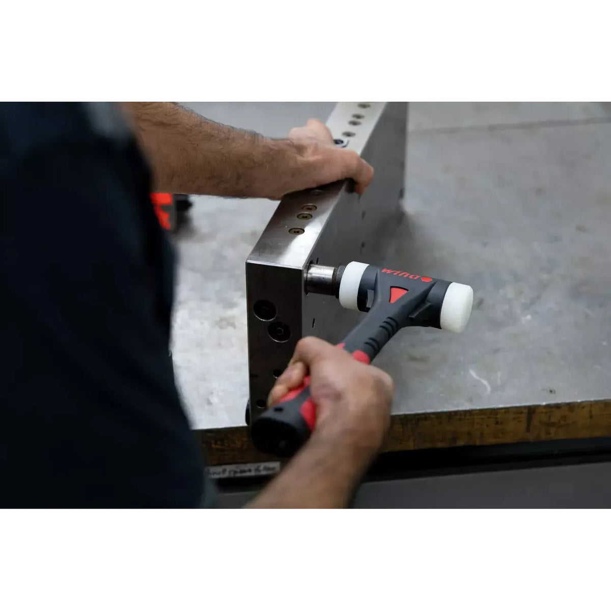A person using a dead blow hammer on a metal block on a workbench.