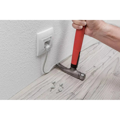 Person using a red-handled electrician's hammer to secure a cable to a wooden floor with clips.