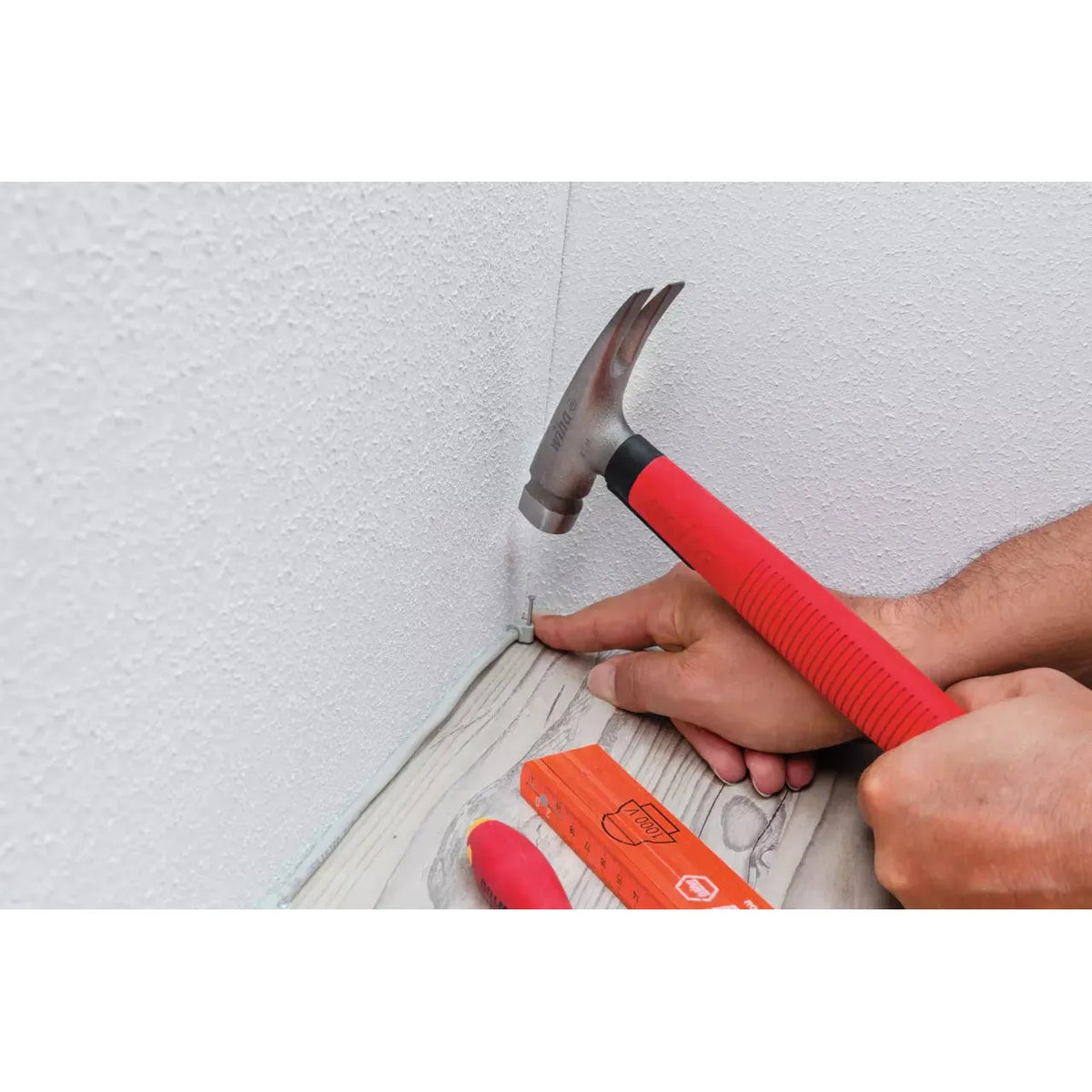Person using a red-handled hammer to secure a cable clip on a baseboard.