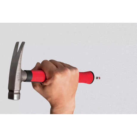 A hand holding a red-handled hammer against a gray wall.