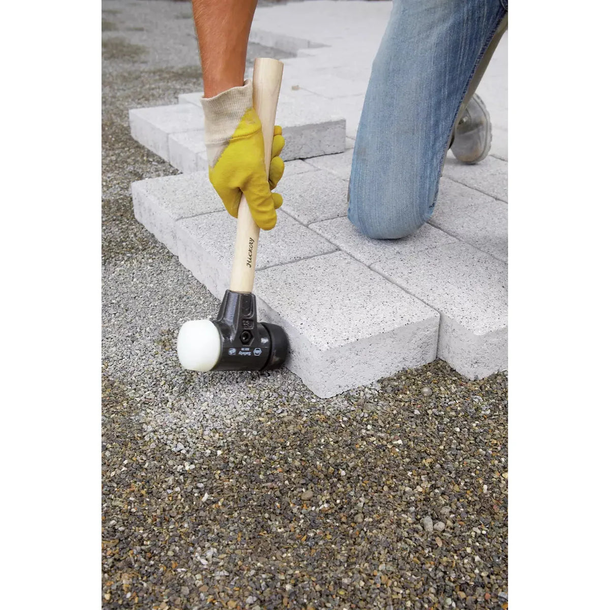 Person using a mallet with a wooden handle to lay paving stones on gravel.