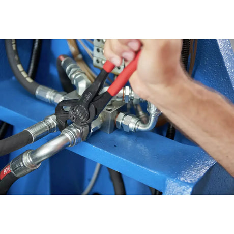 A hand using pliers on a metal fitting with red-handled grips in a mechanical setting.