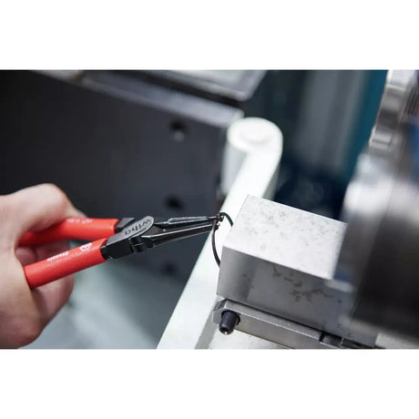 A person uses pliers to remove a retaining ring from a metal block.