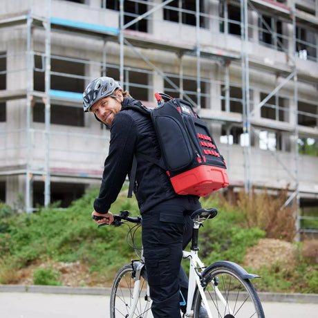A person on a bicycle wearing a helmet and a large black and red backpack in front of a building under construction.