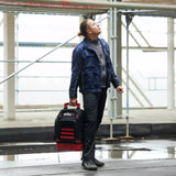 Person in workwear carrying a tool case and looking upwards, construction site environment.