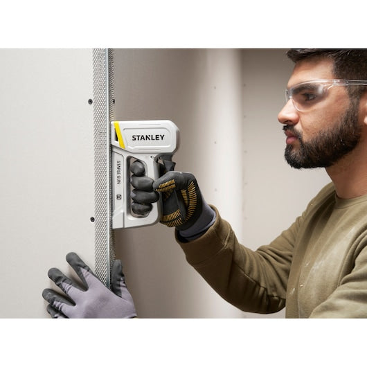 Photo of STANLEY STHT70150-0 staple gun in use, held against drywall by a gloved hand.