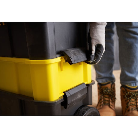 STANLEY STST1-80151 tool box close-up, yellow and black latch, worker with glove and boots visible.